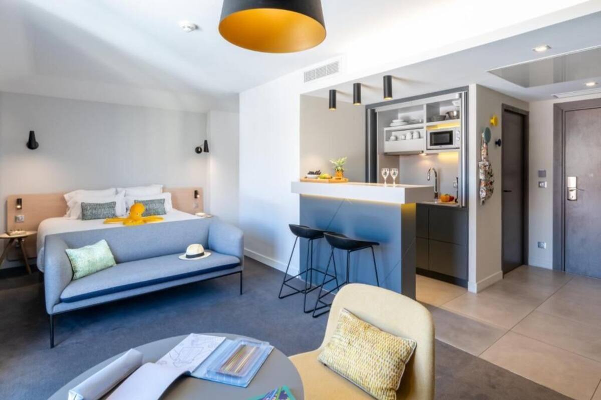 A modern hotel room with a bed, sofa, dining area, and kitchenette.