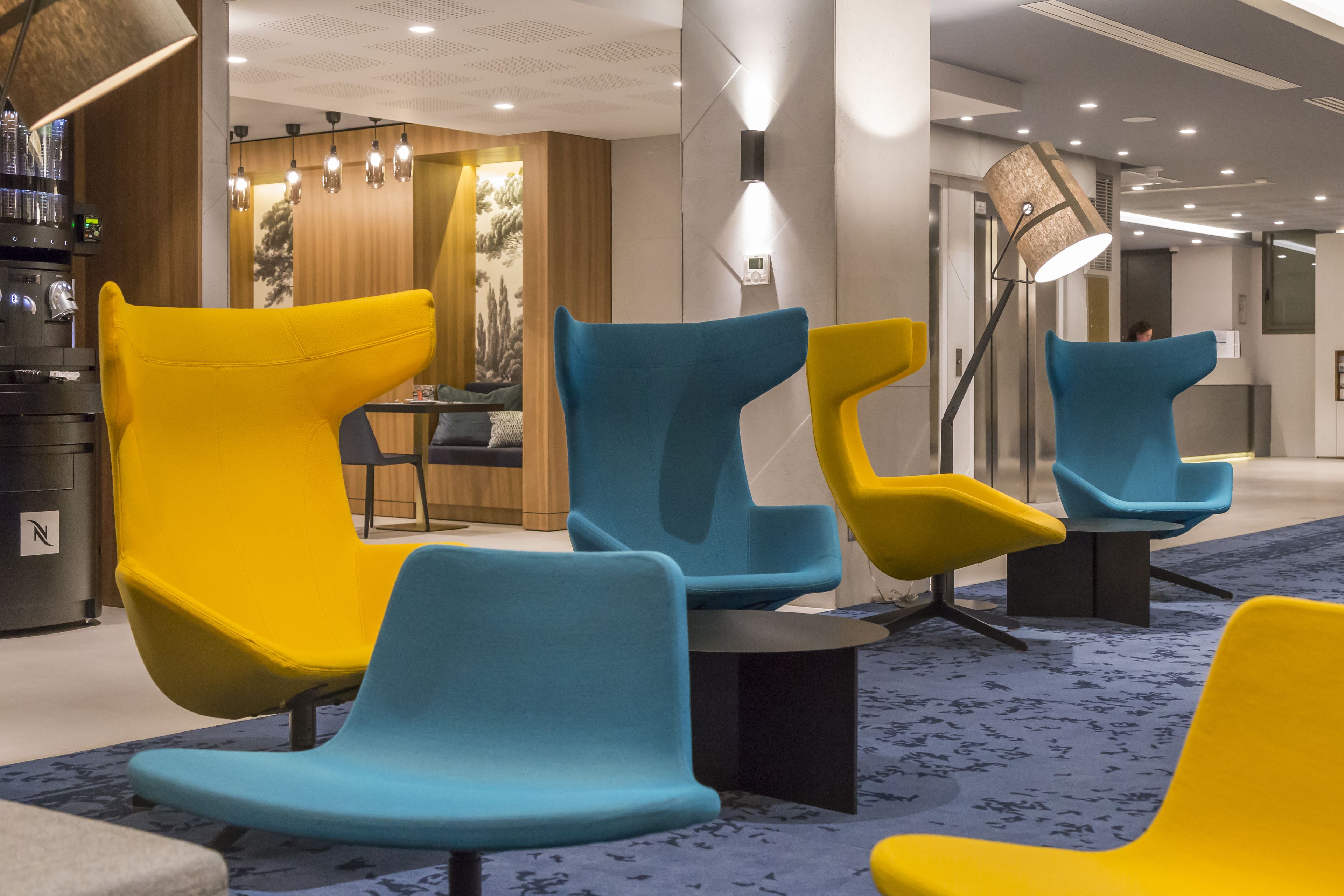 A modern lobby with vibrant yellow and blue chairs, a bar area, and a reception desk.