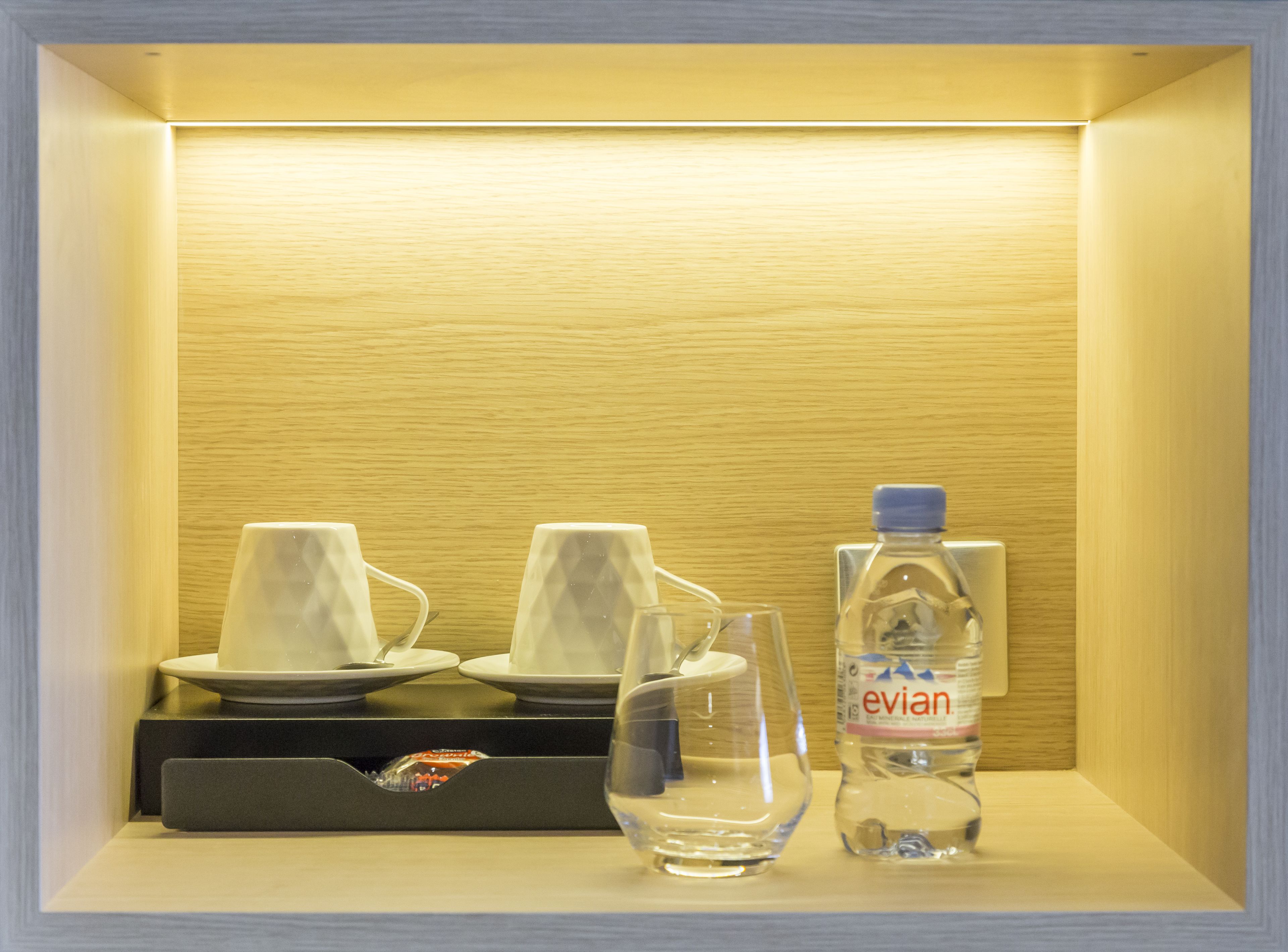 Een netjes ingerichte minibar met een fles Evian-water, twee glazen en een klein bordje met een vork.