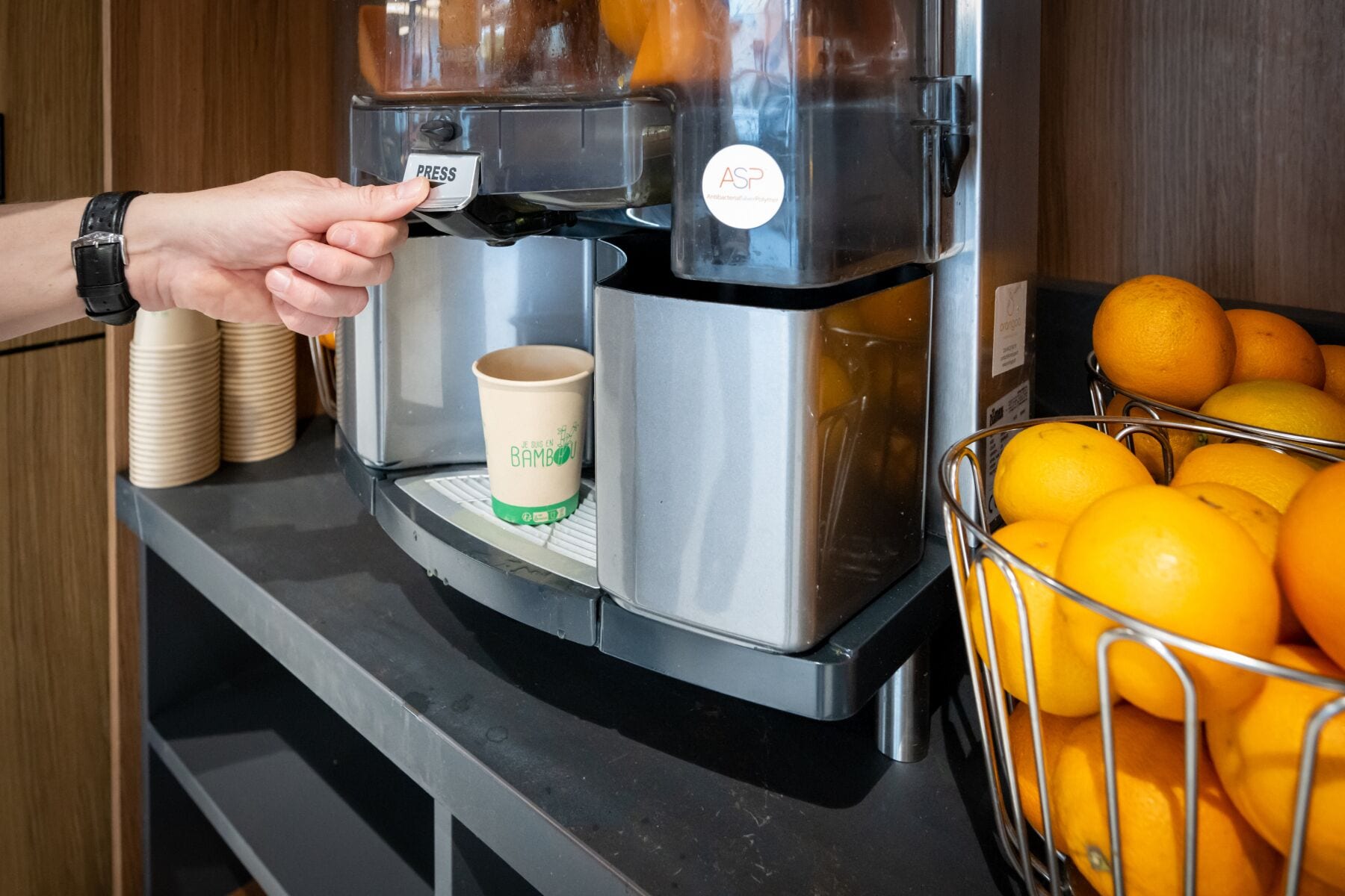 Eine Kaffeemaschine füllt eine Tasse in ein Bambusgefäß, nebenan ein Korb mit Orangen.