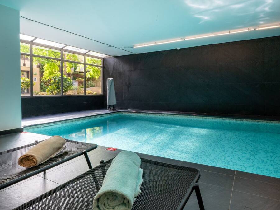 An indoor swimming pool with clear blue water, a dark tiled wall, and a window with a view of greenery outside.