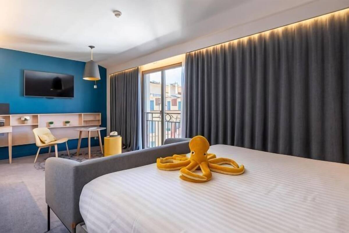 A hotel room with a bed, a desk, a chair, a TV, and a balcony with curtains.