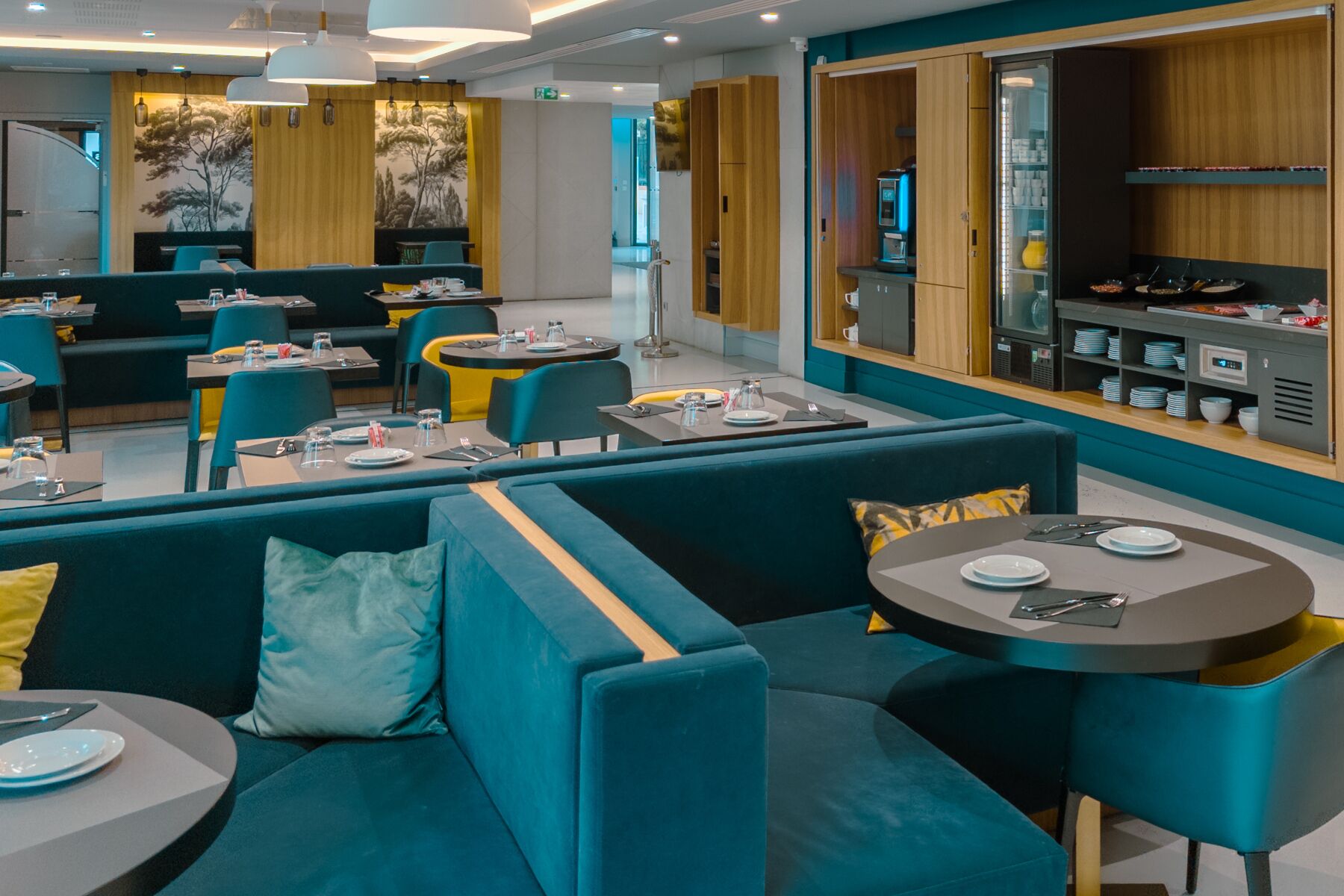 A modern restaurant with teal and yellow seating, a bar area, and a kitchen visible through a glass partition.