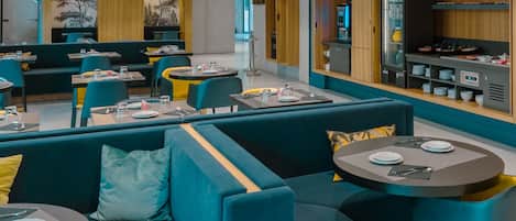 A modern restaurant with teal and yellow seating, a bar area, and a kitchen visible through a glass partition.