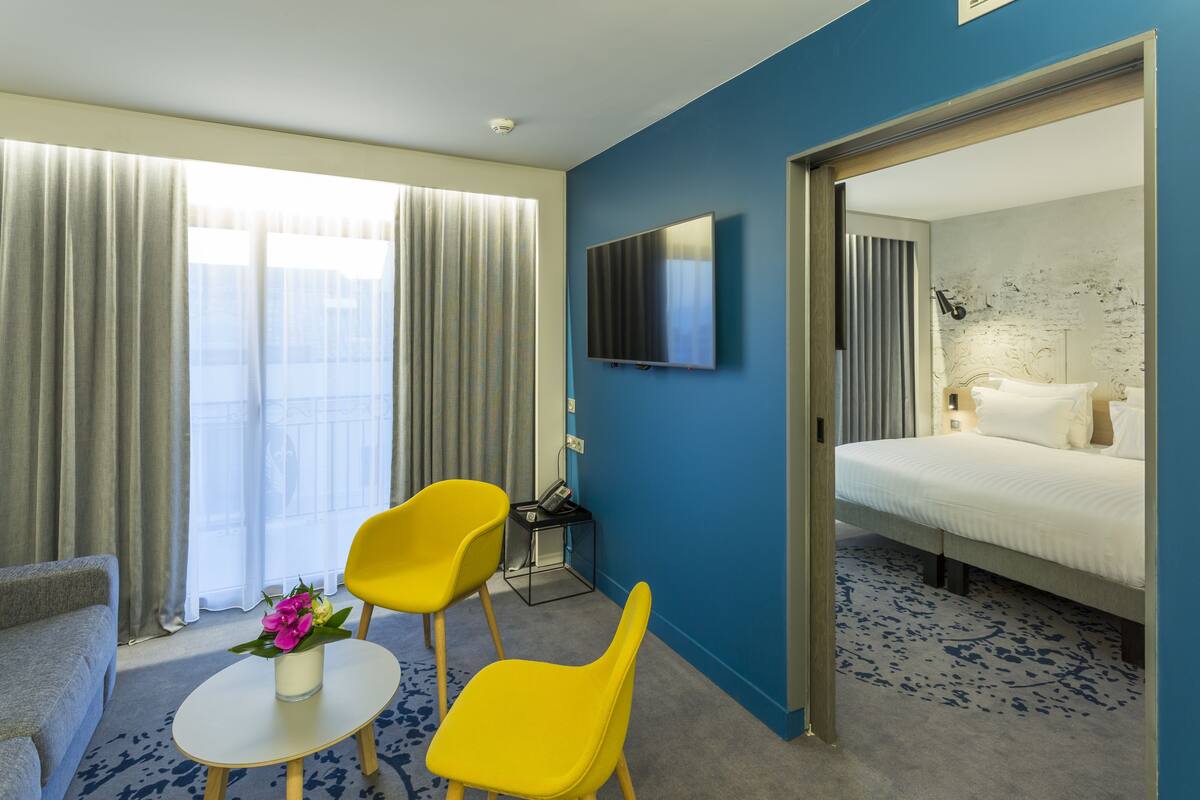 A hotel room with a bed, a TV mounted on the wall, a desk with a chair, and a small table with a vase of flowers.