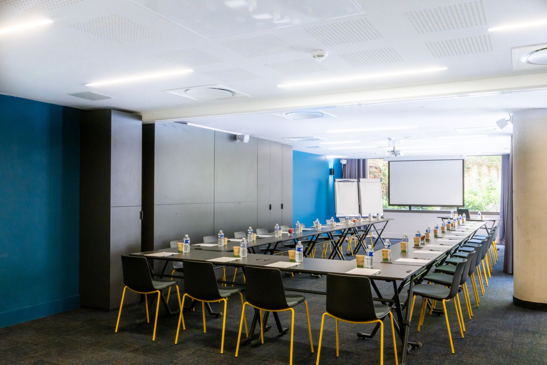 A conference room with a long table, chairs, water bottles, and a projector screen.