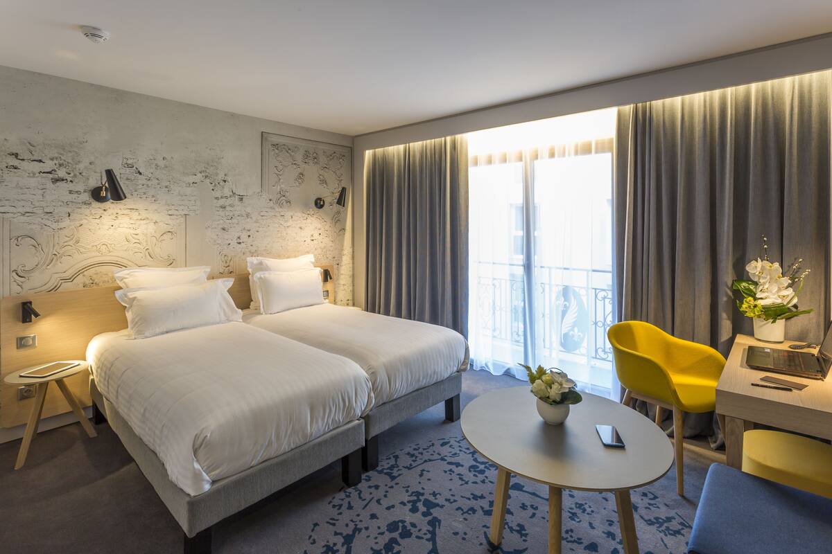 A hotel room with two beds, a desk with a laptop, a yellow chair, and a round table with a vase of flowers.