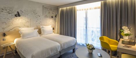 A hotel room with two beds, a desk with a laptop, a yellow chair, and a round table with a vase of flowers.