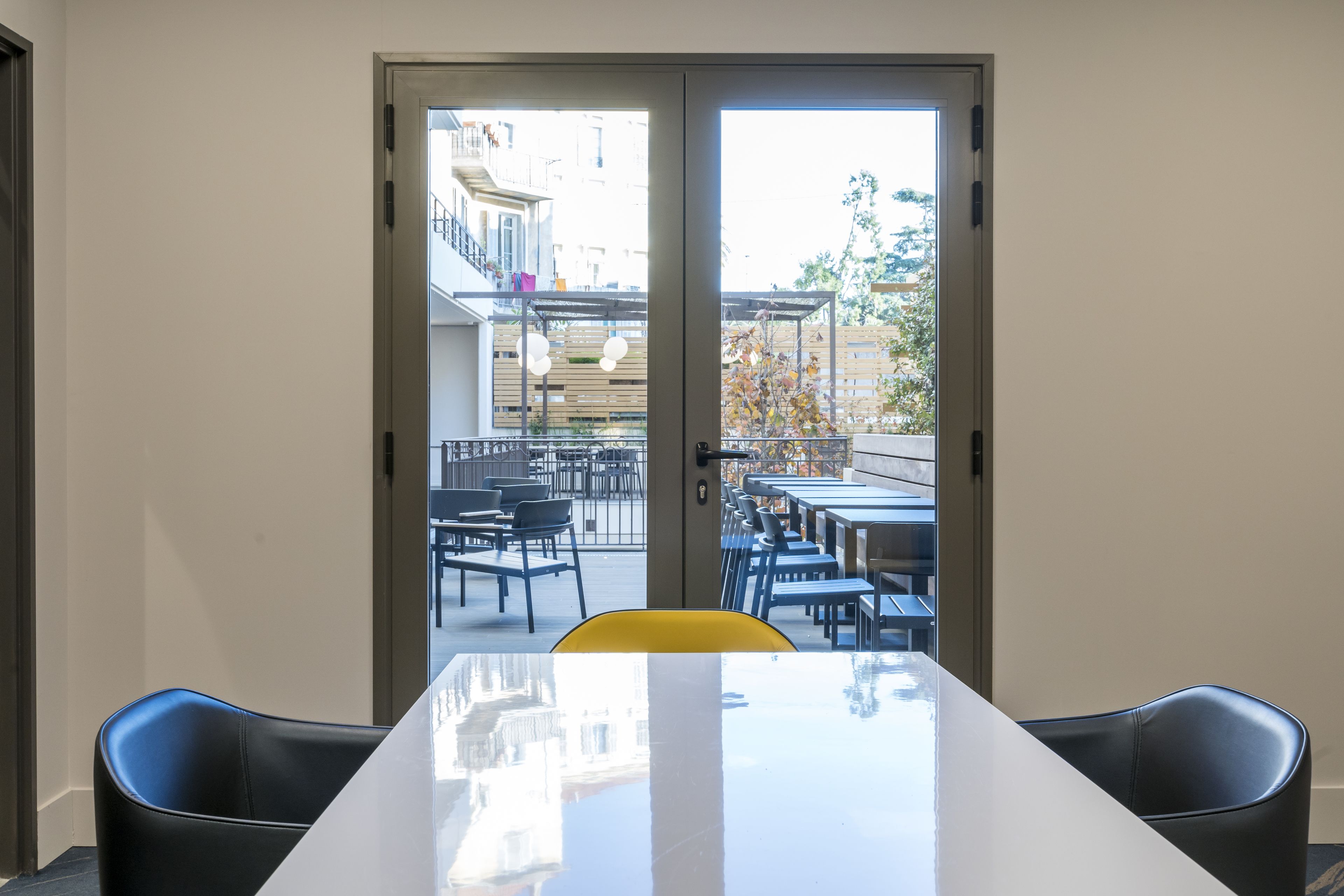 A conference room with a white table and black chairs, a glass door leading to an outdoor seating area with tables and chairs.