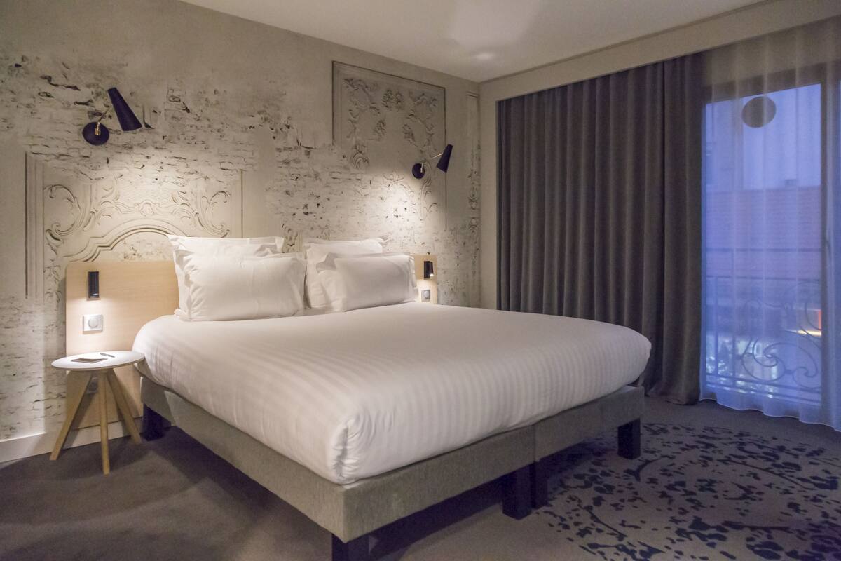 A large bed with white linens, a nightstand with a lamp, and a view of a building through a window.