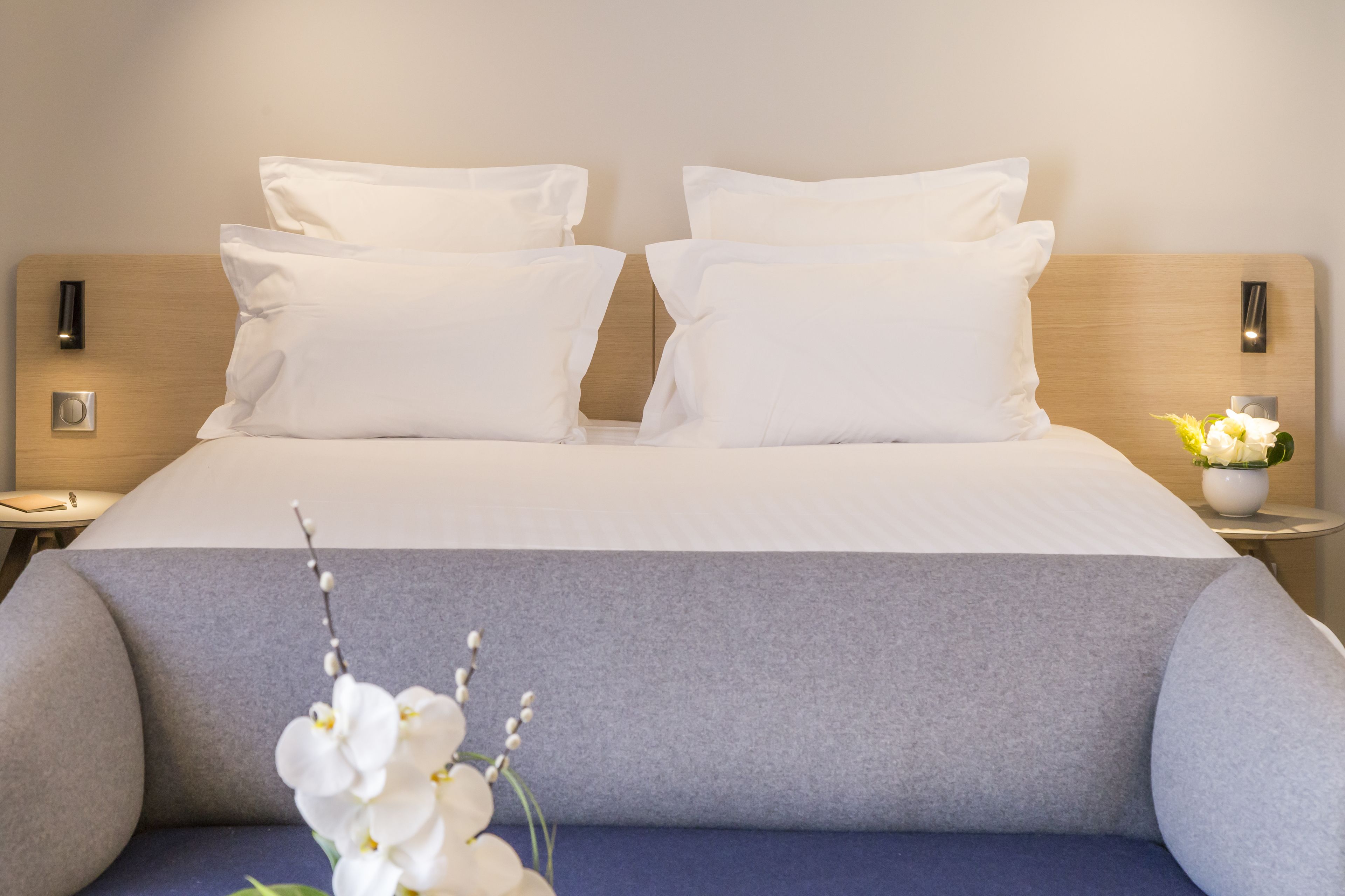 Ein ordentlich bezogenes Bett mit weißen Leinentüchern und zwei Kissen, ein Nachttisch mit einer Vase weißer Blumen sowie eine wandmontierte Leuchte.