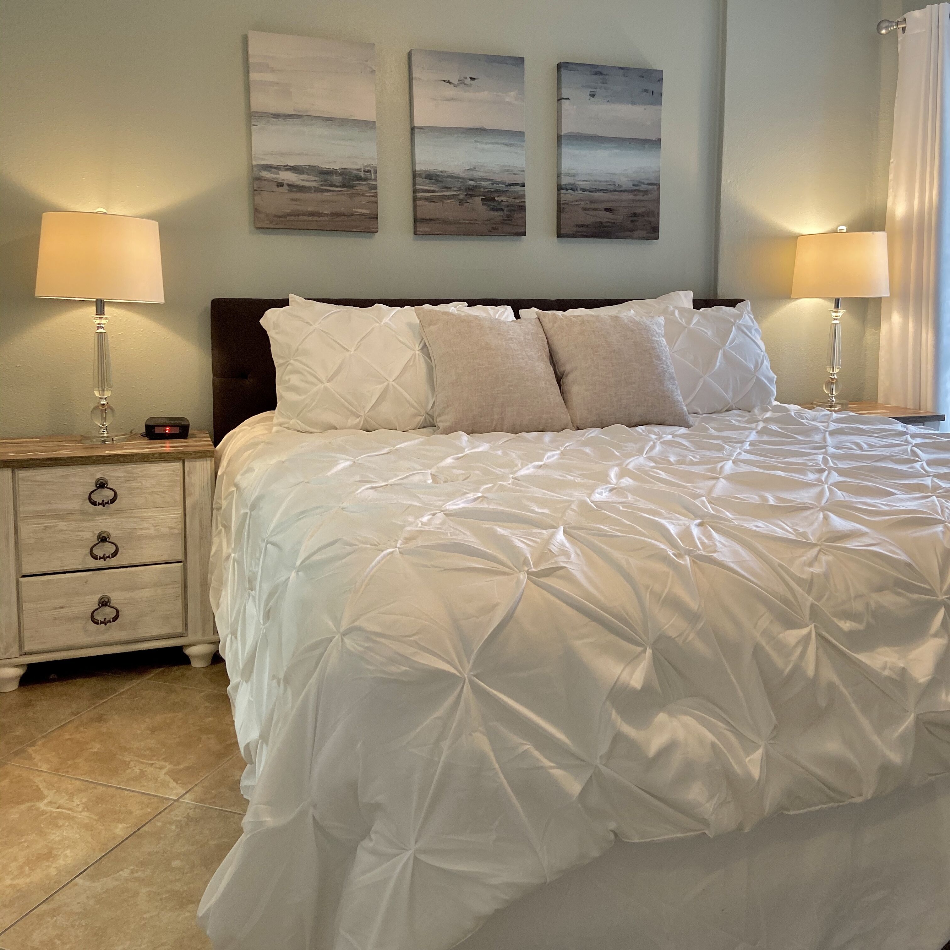 A neatly made bed with a white duvet, two bedside tables with lamps, and three wall-mounted paintings.