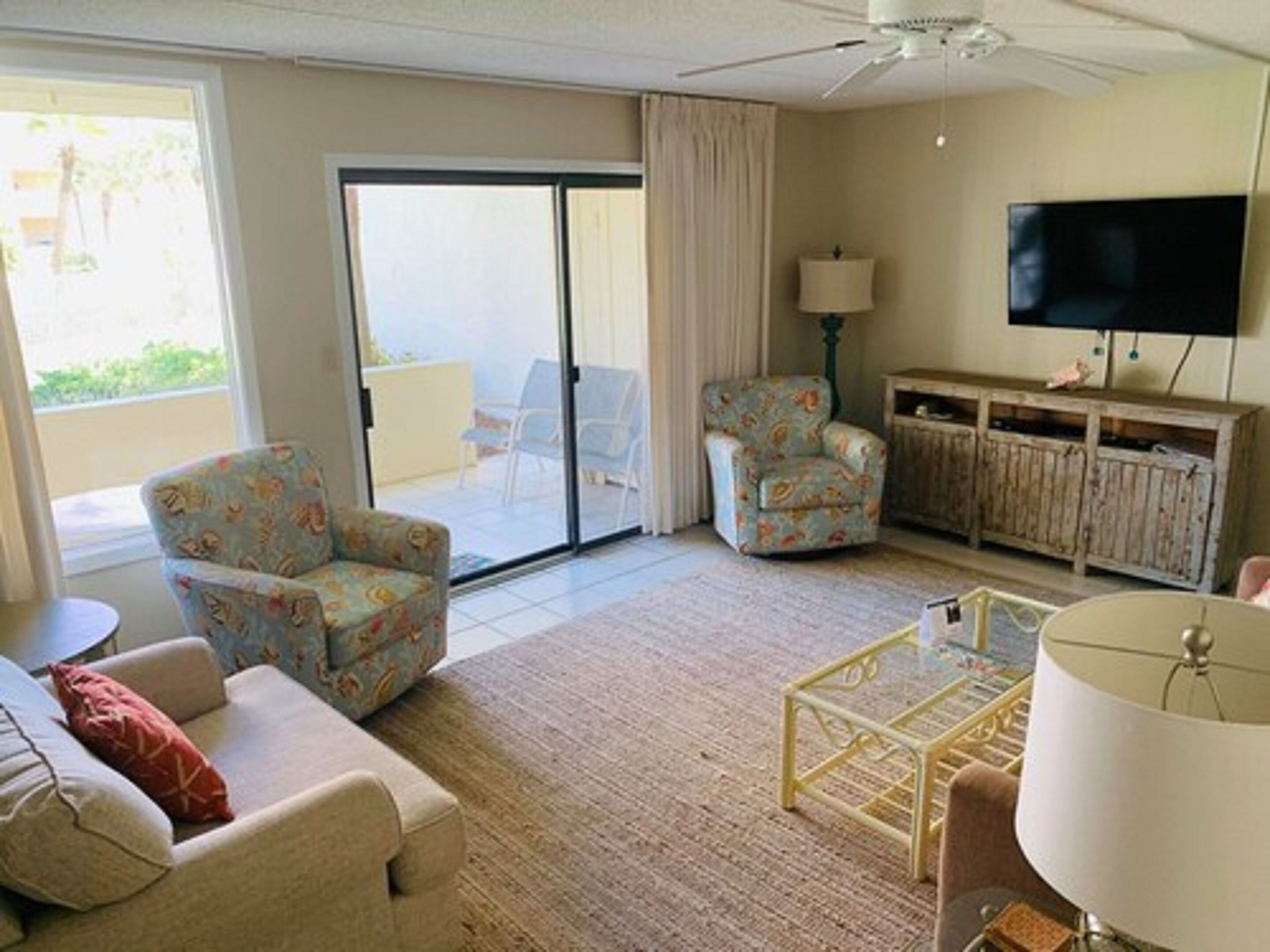 A living room with a sofa, two patterned armchairs, a coffee table, a TV, and a sliding glass door leading to a balcony.