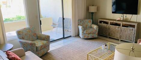 A living room with a sofa, two patterned armchairs, a coffee table, a TV, and a sliding glass door leading to a balcony.