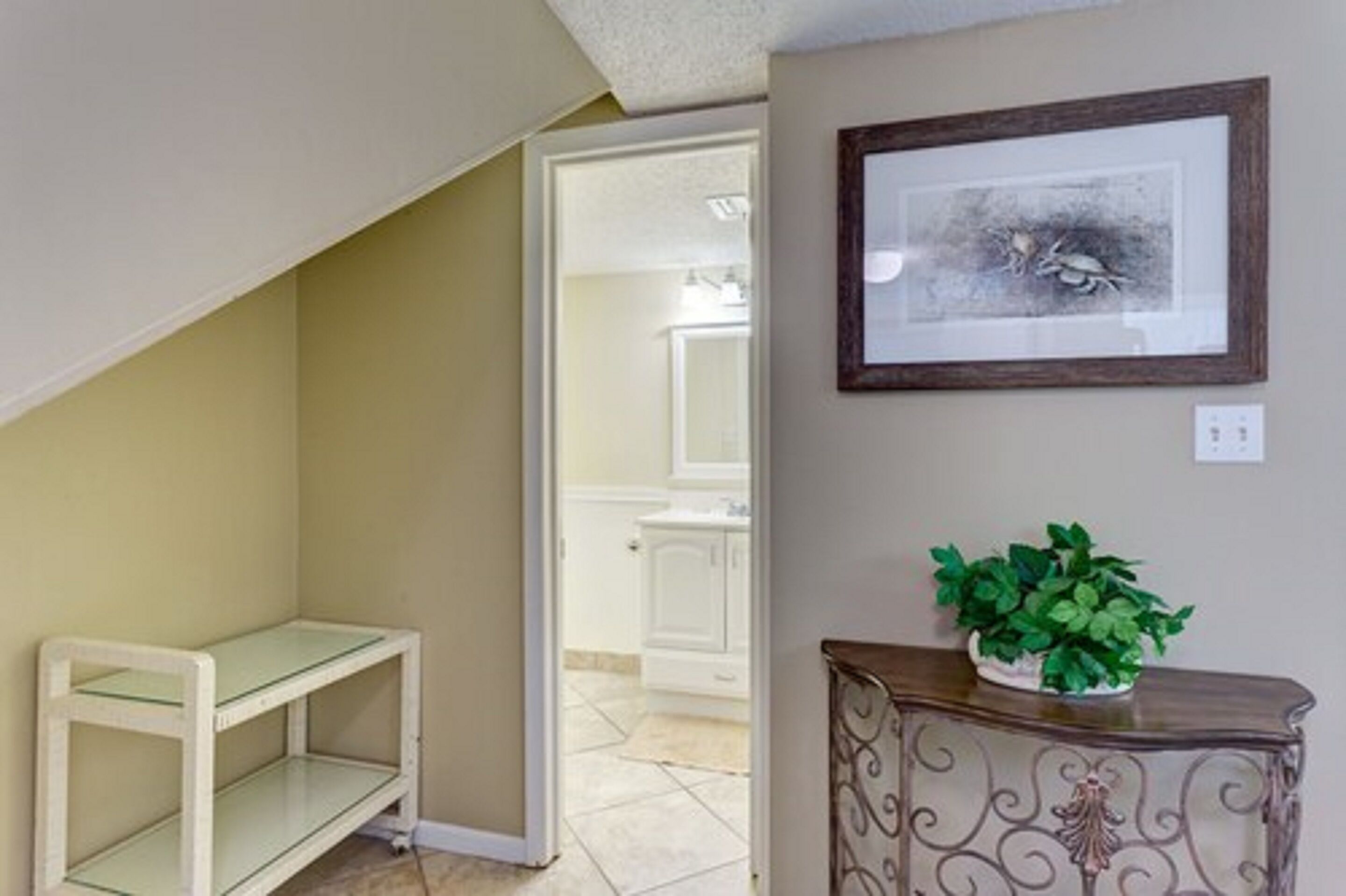 Un couloir avec une porte blanche donnant sur une salle de bain, un tableau encadré accroché au mur et une petite table sur laquelle est posée une plante.