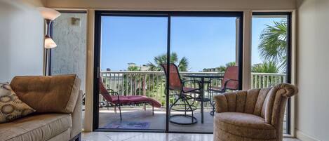 Una sala de estar con sofá, sillón y mesa de comedor con sillas. Hay una puerta corrediza de vidrio que da a un patio al aire libre con vista a palmeras y edificios.