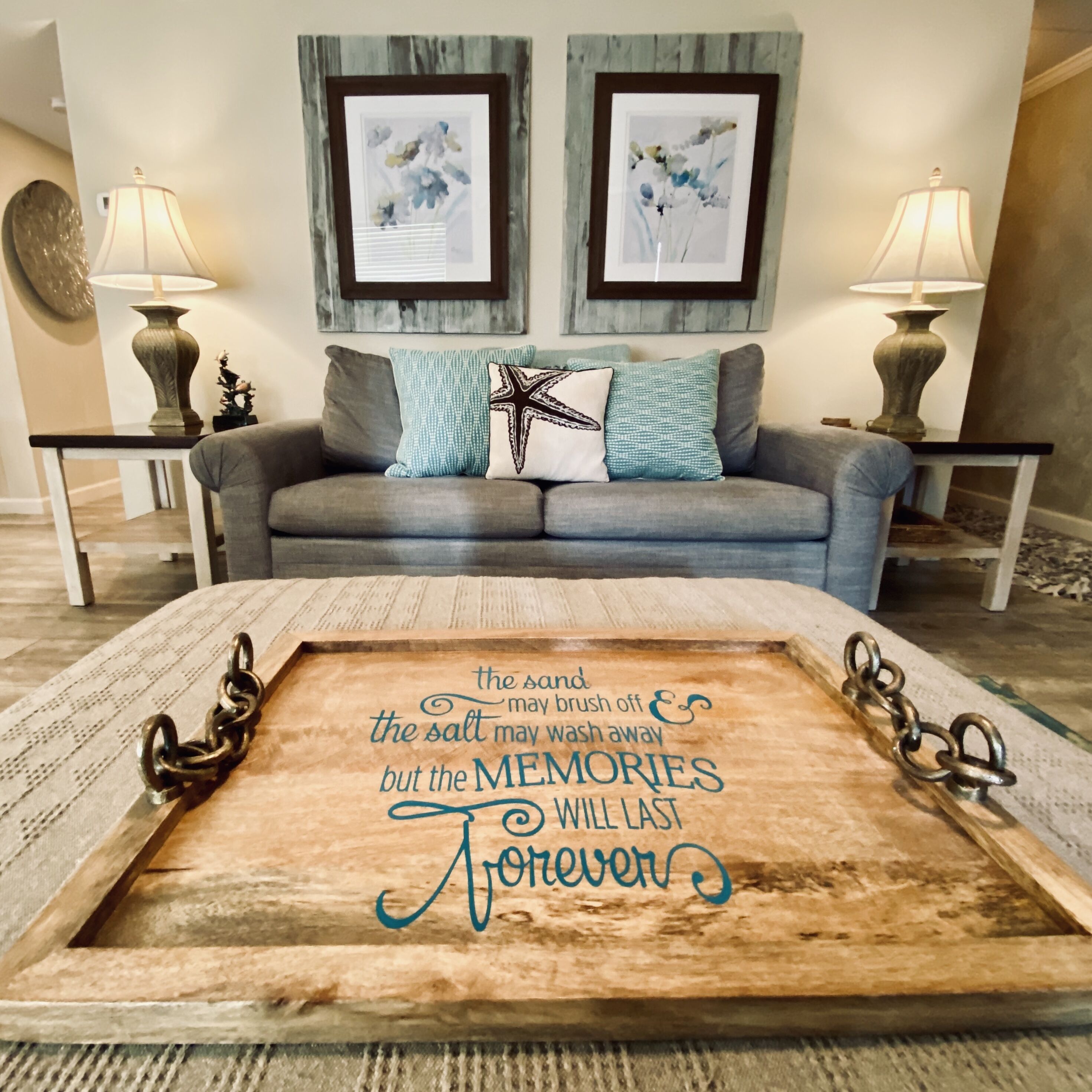A living room with a wooden coffee table displaying a sentimental quote, a grey sofa with patterned cushions, and two framed artworks above the sofa.