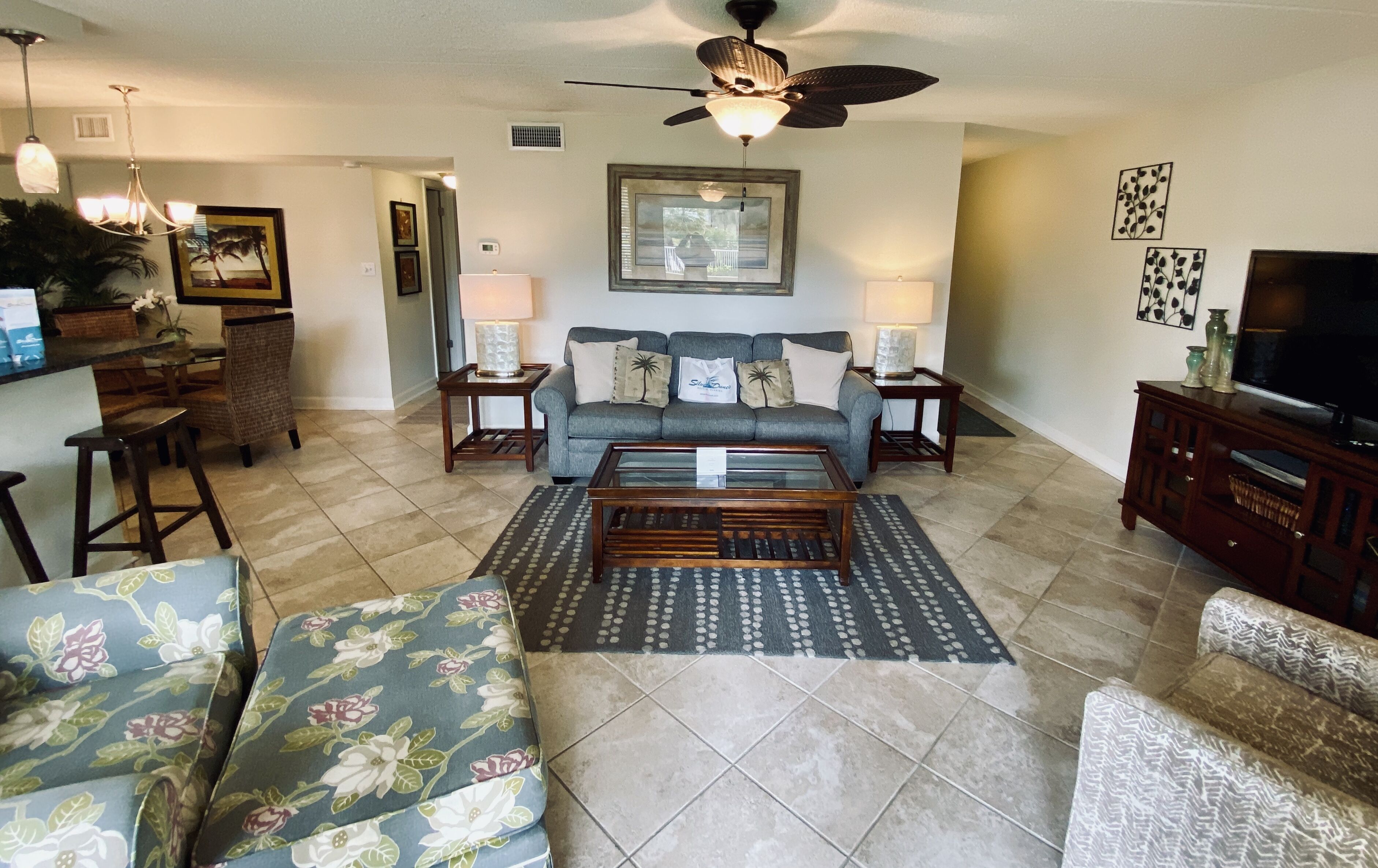 A living room with a sofa, coffee table, and patterned rug. There is a television, a ceiling fan, and a framed picture on the wall.