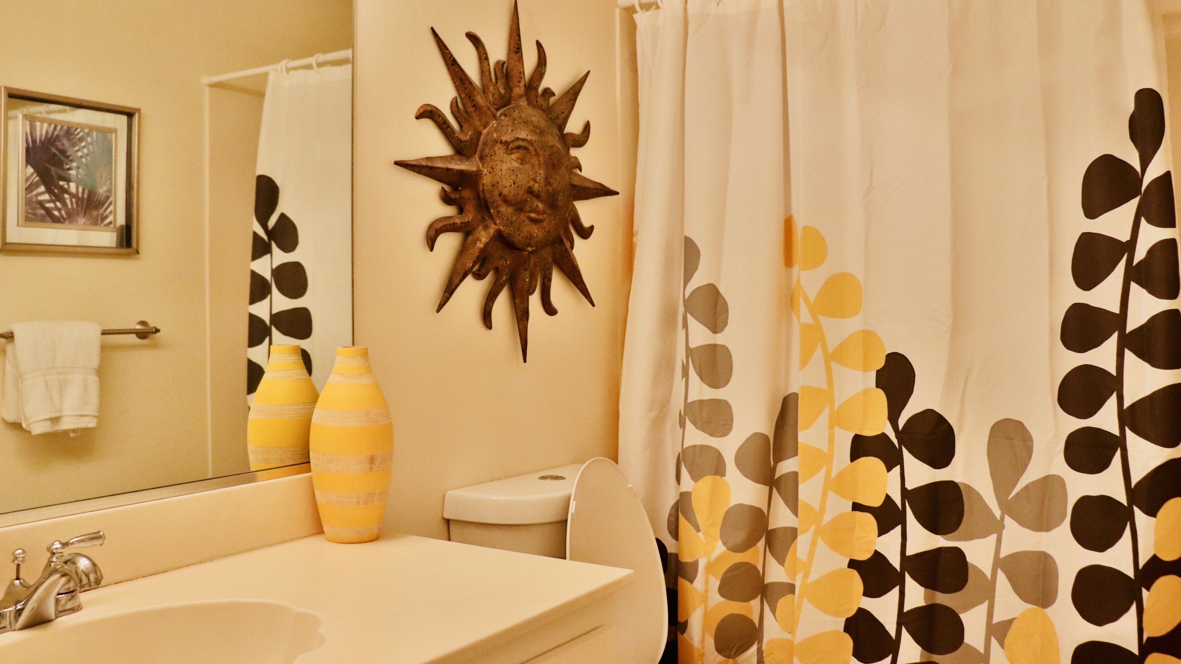 Un baño con un lavamanos, dos jarrones a rayas, un adorno de sol y una cortina de ducha con estampado de hojas.