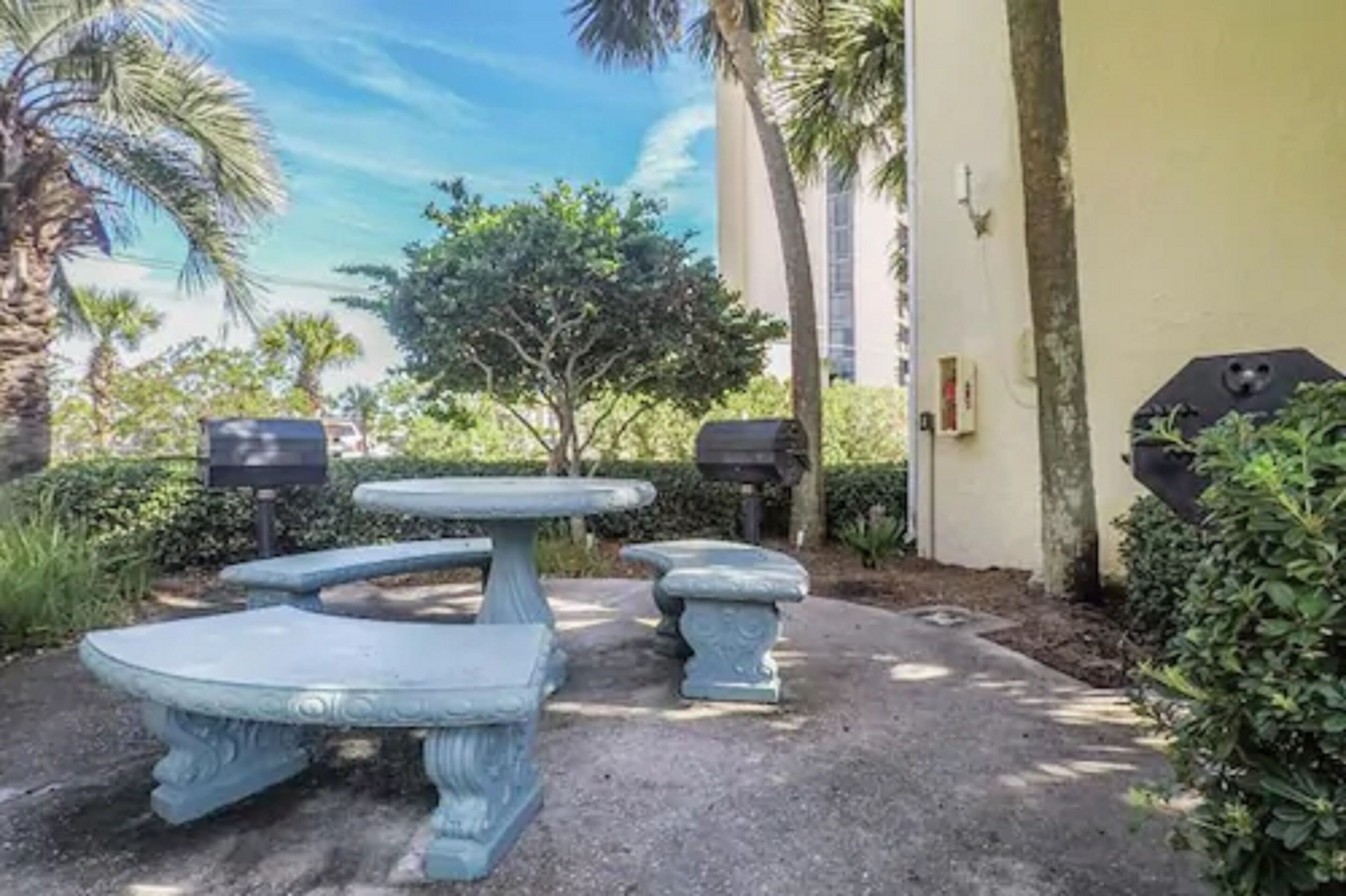 An outdoor seating area with a table and benches, surrounded by greenery and a building in the background.