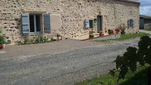Un bâtiment en pierre avec deux fenêtres et une porte, entouré de plantes en pot et bordé d’une allée gravillonnée.