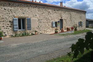A stone building with two windows and a door, surrounded by potted plants and a gravel driveway.