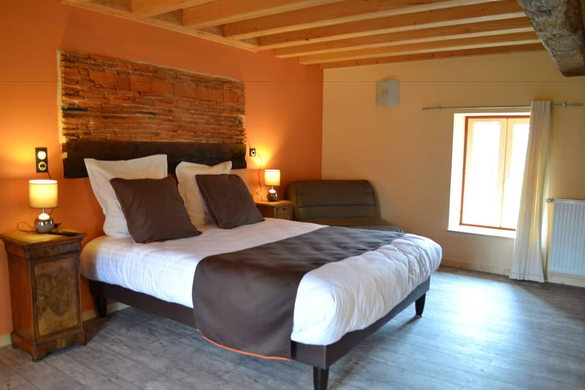 Quarto com parede de tijolos decorativa, uma cama grande com colcha marrom, duas luminárias de cabeceira e uma janela com cortinas.