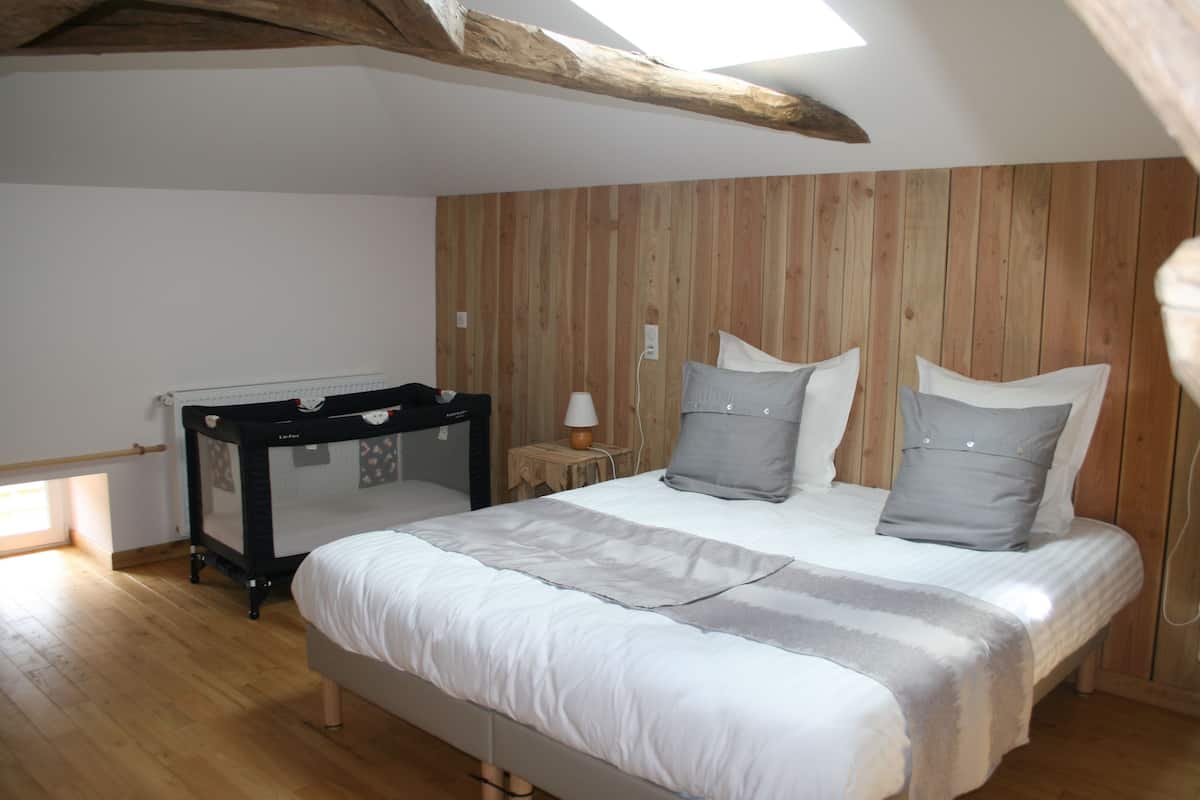 Quarto com cabeceira de madeira, cama com roupa de cama cinza, mesa de cabeceira com abajur e berço no canto.