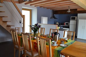 A dining area with wooden chairs and a table set for a meal, a kitchen in the background, and a staircase leading upstairs.