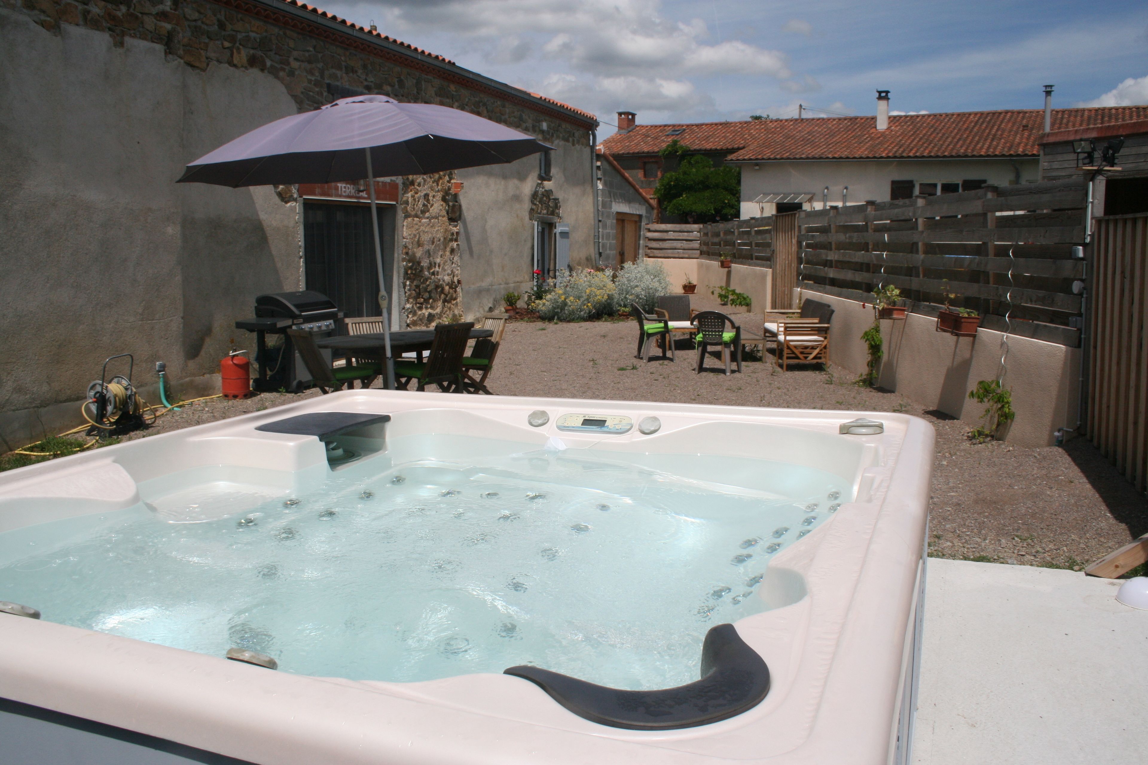 Un jacuzzi installé dans un jardin, avec un espace patio, un barbecue et une clôture en bois.