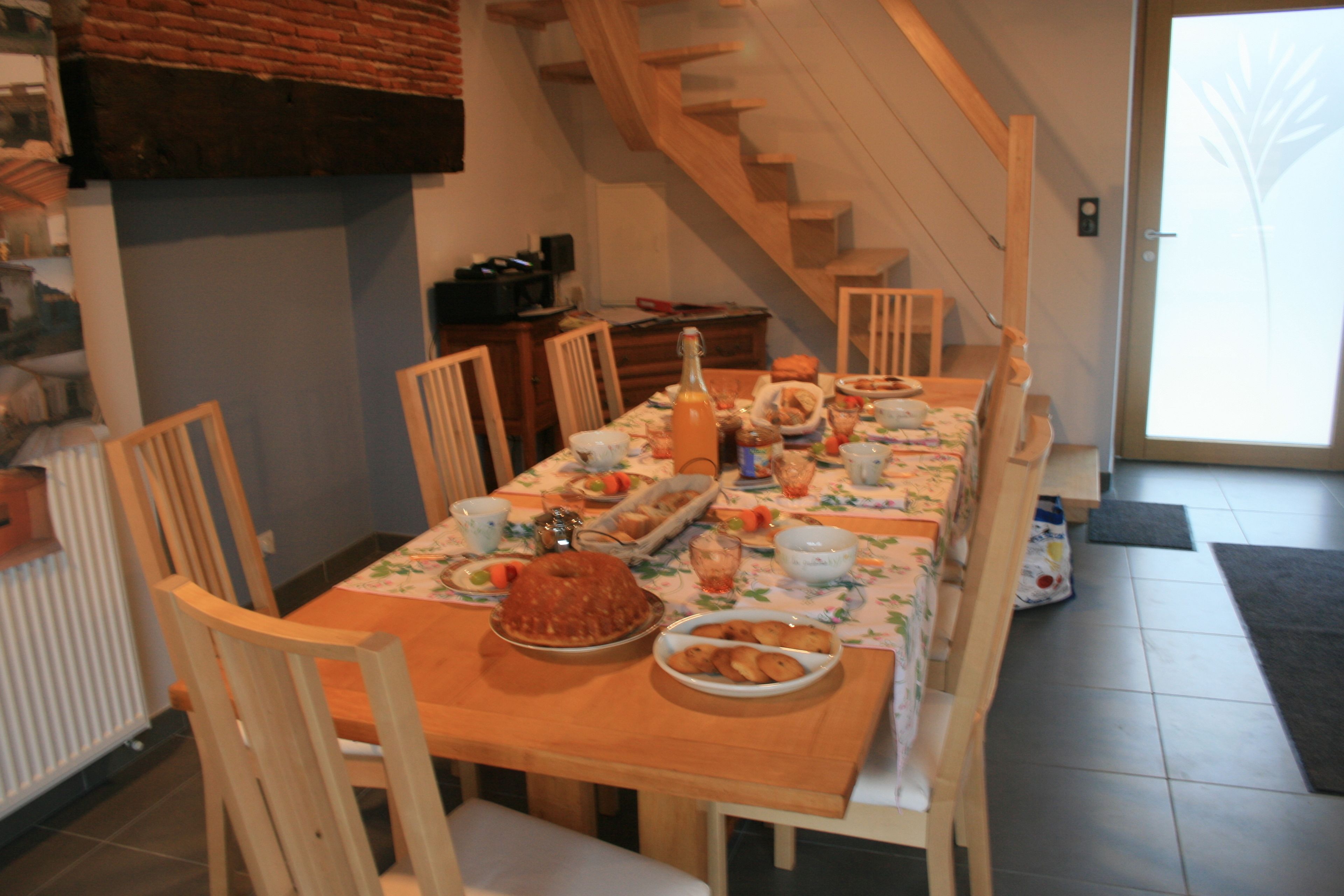 A dining area with a wooden table set for a meal, surrounded by chairs. A staircase is visible in the background.