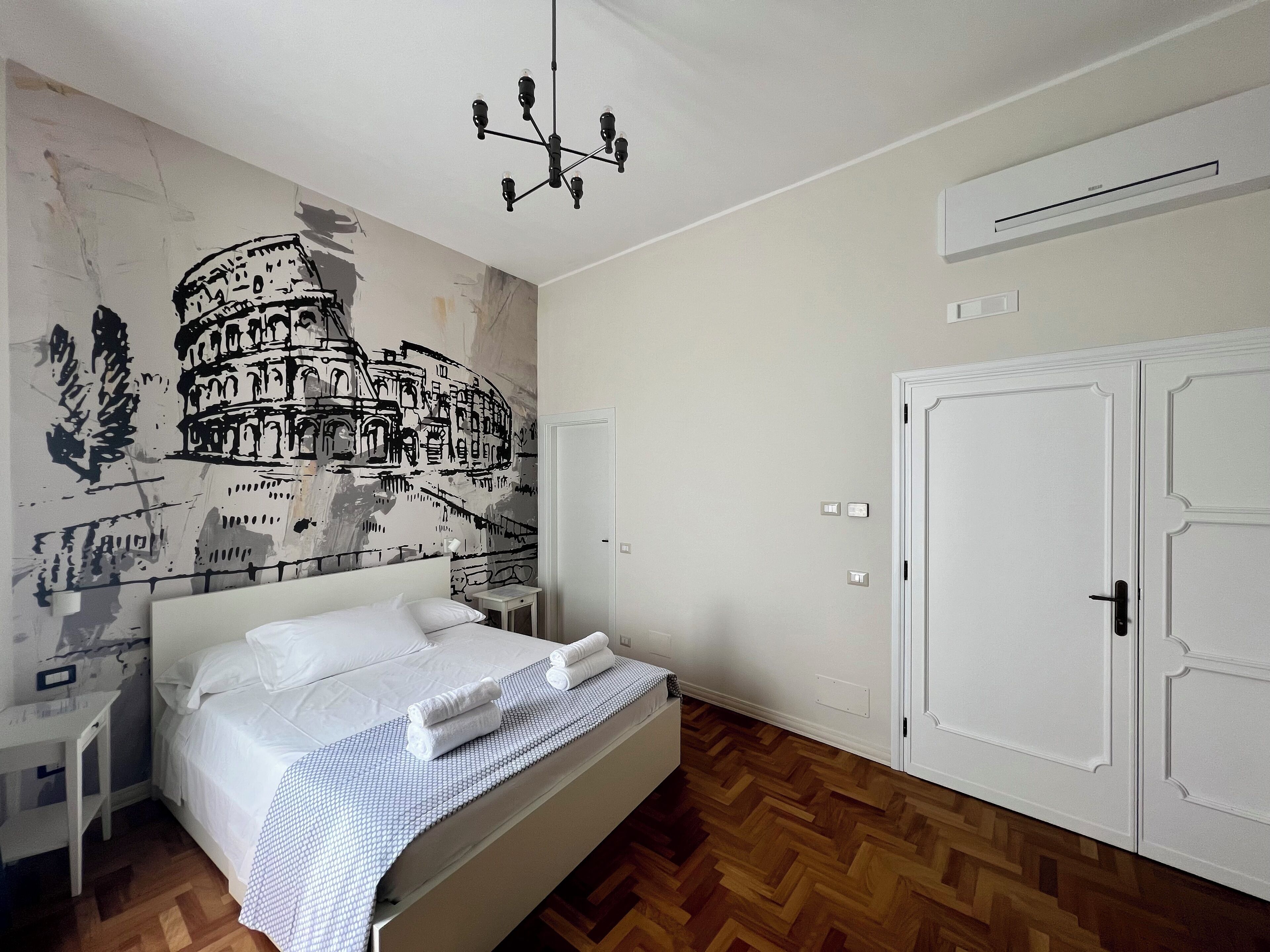 A hotel room with a bed, a nightstand, a wall mural of the Colosseum, a chandelier, and a wardrobe.