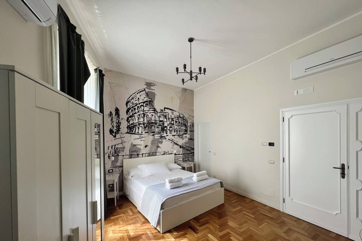 Een slaapkamer met een bed, een houten visgraatvloer, een muurschildering van het Colosseum en een witte kledingkast.