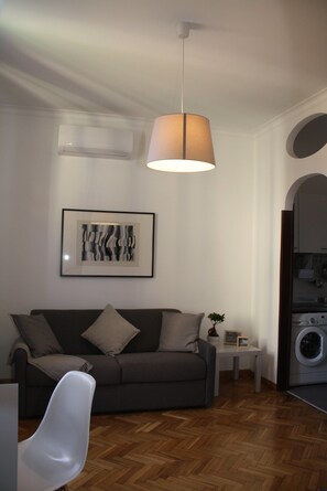 A modern living room with a grey sofa, a white chair, a hanging lamp, a framed artwork, a small table with a plant, and a washing machine in the background.