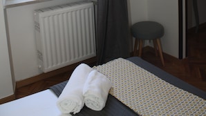 Ein Schlafzimmer mit Bett, Heizkörper, einem fenster mit Vorhängen, einem Hocker und einem gerahmten Bild an der Wand.