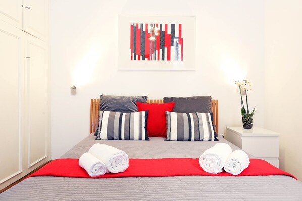 A neatly made bed with striped pillows, a red throw blanket, and rolled towels.