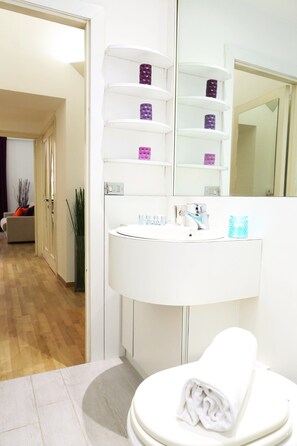 A modern bathroom with a white sink, mirror, and toilet. There are shelves above the sink with toiletries. - Home2Rome - Trastevere Natale (Rome)