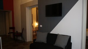 A living room with a black sofa, two grey cushions, a flat-screen TV mounted on the wall, and a dining area with a table and chairs. - Home2Rome - Trastevere Del Grande (Rome)