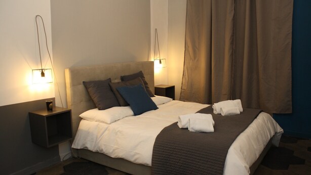 A neatly made bed with white and brown bedding, two bedside tables with lamps, and a large window with curtains. - Home2Rome - Trastevere Del Grande (Rome)