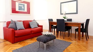 A modern living room with a red sofa, grey cushions, a grey ottoman, a dining table with chairs, and a large mirror on the wall. - Home2Rome - Trastevere Roma Libera (Rome)