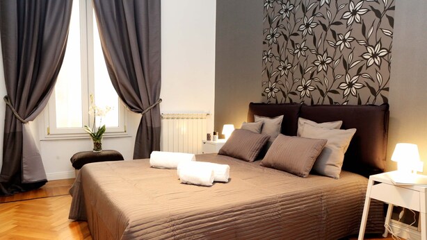 A neatly made bed with a brown bedspread, two pillows, and two rolled towels. There is a nightstand with a lamp, a radiator, and a wall with a floral pattern. - Home2Rome - Trastevere Roma Libera (Rome)