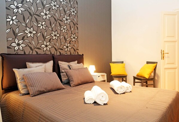 A neatly made bed with a brown quilt, two rolled towels, and a floral-patterned headboard. - Home2Rome - Trastevere Roma Libera (Rome)