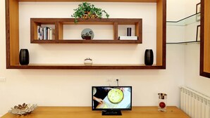 A living room with a wooden TV stand, a flat-screen TV, a bookshelf with books and decorative items, and a potted plant. - Home2Rome - Trastevere Roma Libera (Rome)