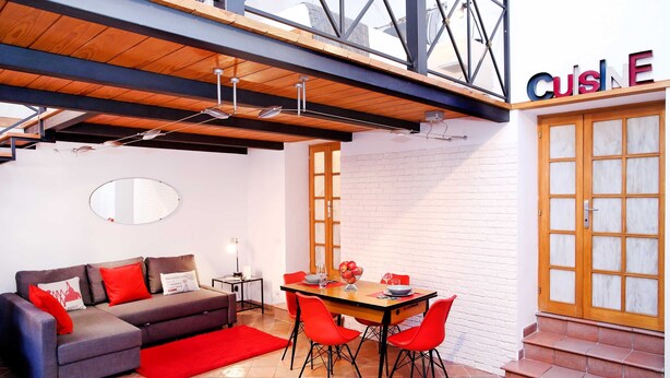 A modern living area with a wooden ceiling, a dining table with red chairs, a grey sofa with red cushions, and a large window with a view of the city. - Home2Rome - Trastevere Galletti (Rome)