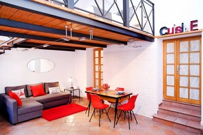 A modern living area with a wooden ceiling, a dining table with red chairs, a grey sofa with red cushions, and a large window with a view of the city.
