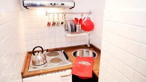 A kitchen with a stove, kettle, and sink. There is a red towel hanging on the oven handle and a shelf with kitchen utensils. - Home2Rome - Trastevere Galletti (Rome)