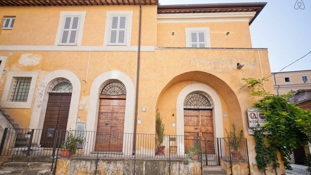 A building with three arched doorways, a yellow facade, and a small garden area with potted plants.