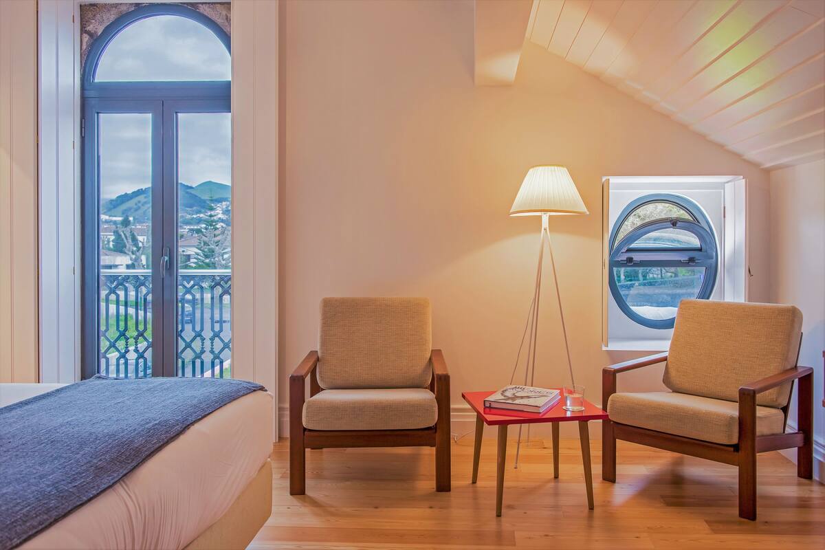 A room with a bed, two armchairs, a small table with a book and glass, a lamp, and a window with a view of hills and a bridge.