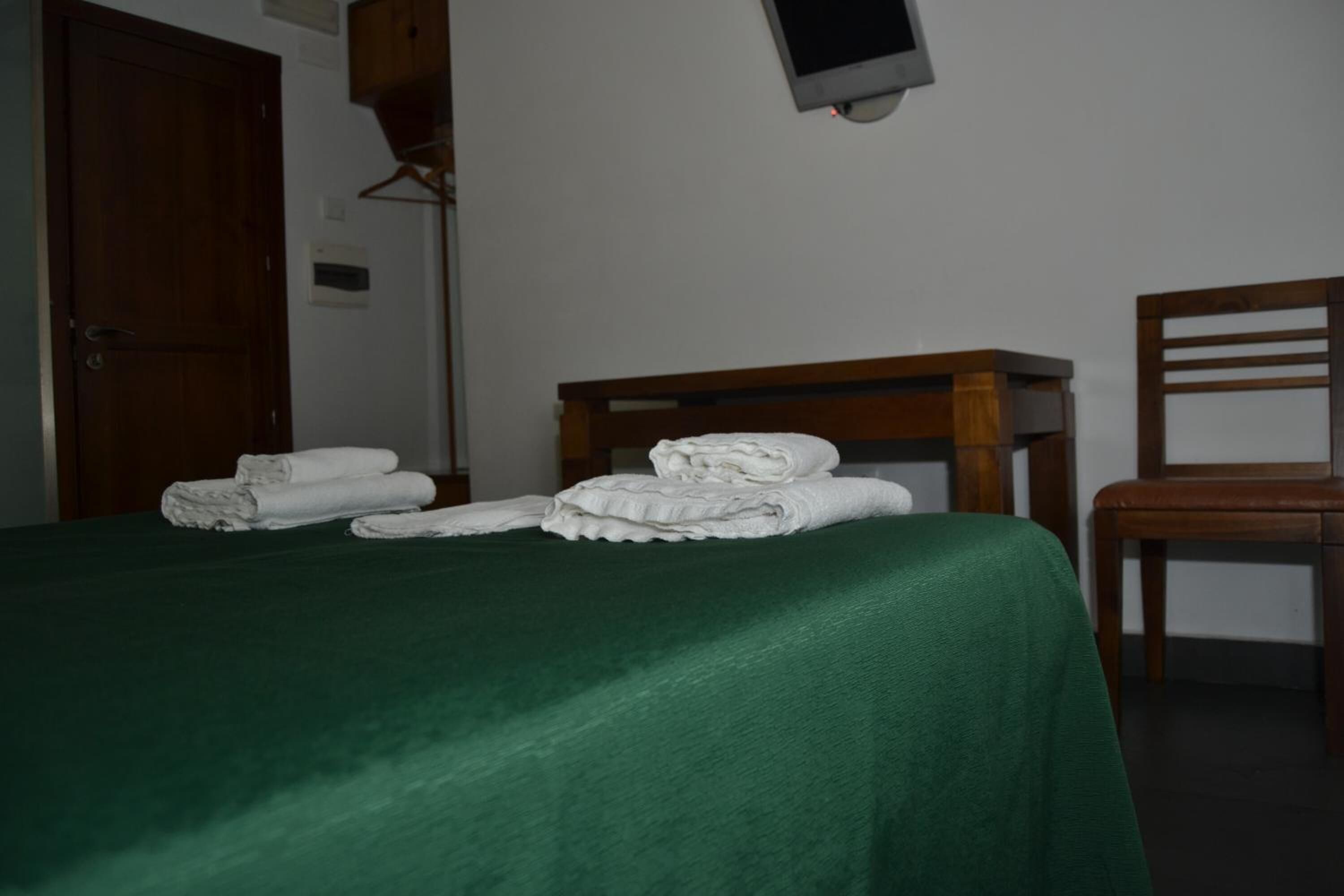 A hotel room with a bed covered in a green sheet, a wooden chair, and a wooden nightstand with a television.