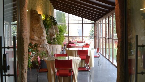 Un comedor interior con mesas y sillas, un pilar de piedra, y vistas a través de amplios ventanales.